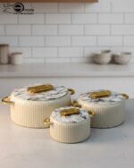 A professional studio shot of a set of three graduated-size insulated hotpots with gold handles, featuring a ribbed cream or grey body and decorative marble or wood-patterned lids, displayed on a kitchen countertop.