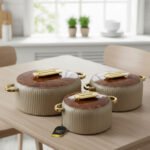 A professional studio shot of a set of three graduated-size insulated hotpots with gold handles, featuring a ribbed cream or grey body and decorative marble or wood-patterned lids, displayed on a kitchen countertop.