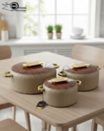 A professional studio shot of a set of three graduated-size insulated hotpots with gold handles, featuring a ribbed cream or grey body and decorative marble or wood-patterned lids, displayed on a kitchen countertop.