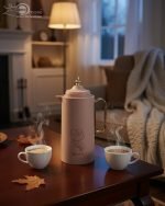 A professional display of three 1-liter Feenik vacuum thermoses in Black, White, and Coffee colors. Each flask features a gold-accented handle, a decorative gold floral print, and a push-to-pour lid, set against a clean kitchen background.