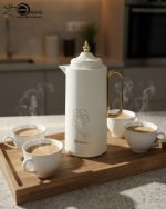 A professional display of three 1-liter Feenik vacuum thermoses in Black, White, and Coffee colors. Each flask features a gold-accented handle, a decorative gold floral print, and a push-to-pour lid, set against a clean kitchen background.