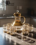 Elegant 7-piece glass beverage set with gold accents including a decanter and six tumblers, shown on a table setting and being used to pour a golden drink.