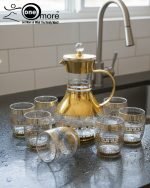 Elegant 7-piece glass beverage set with gold accents including a decanter and six tumblers, shown on a table setting and being used to pour a golden drink.
