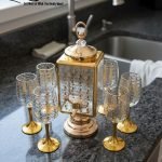 Fancy gold-tone beverage dispenser set with six matching stemmed goblets. The dispenser and glasses have a clear body overlaid with a delicate, scrolling gold floral pattern, and the goblets have solid gold bases.