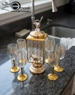 Fancy gold-tone beverage dispenser set with six matching stemmed goblets. The dispenser and glasses have a clear body overlaid with a delicate, scrolling gold floral pattern, and the goblets have solid gold bases.