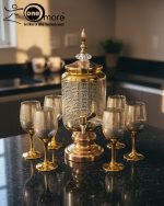 Fancy gold-tone beverage dispenser set with six matching stemmed goblets. The dispenser and glasses have a clear body overlaid with a delicate, scrolling gold floral pattern, and the goblets have solid gold bases.