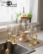 Fancy gold-tone beverage dispenser set with six matching stemmed goblets. The dispenser and glasses have a clear body overlaid with a delicate, scrolling gold floral pattern, and the goblets have solid gold bases.