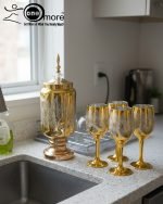 Fancy gold-tone beverage dispenser set with six matching stemmed goblets. The dispenser and glasses have a clear body overlaid with a delicate, scrolling gold floral pattern, and the goblets have solid gold bases.