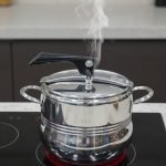 One More stainless steel pressure cooker releasing steam on an induction or ceramic electric cooktop.