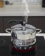 One More stainless steel pressure cooker releasing steam on an induction or ceramic electric cooktop.
