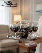 7-piece glass water set, including a clear glass jug/pitcher with white lid and six stemmed wine glasses, all featuring an elegant gold floral design, shown serving beverages in a home setting.