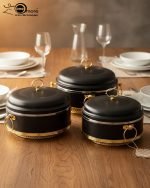 Set of three black insulated hot pot casseroles with gold trim and loop handles, sitting on a wooden dining table with a formal place setting.