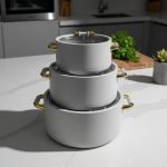 A stacked set of three premium insulated hot pots (1L, 2L, 3.5L) in a crisp white color with decorative gold-toned handles, shown on a marble kitchen countertop. The set features clear acrylic lids and is marketed for keeping food warm.