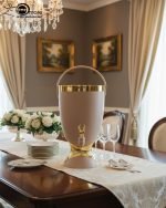 Insulated, stylish beverage dispenser (water cooler) with gold accents on base and spigot, shown in white, green, and chocolate/taupe colors, used for serving drinks on a dining table or kitchen counter.
