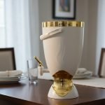 Insulated, stylish beverage dispenser (water cooler) with gold accents on base and spigot, shown in white, green, and chocolate/taupe colors, used for serving drinks on a dining table or kitchen counter.
