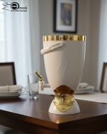 Insulated, stylish beverage dispenser (water cooler) with gold accents on base and spigot, shown in white, green, and chocolate/taupe colors, used for serving drinks on a dining table or kitchen counter.