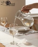 7-piece glass water set, including a clear glass jug/pitcher with white lid and six stemmed wine glasses, all featuring an elegant gold floral design, shown serving beverages in a home setting.