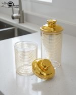 A set of three ribbed acrylic storage jars in different sizes, some with clear lids and some with shiny gold lids, filled with various nuts and snacks, displayed on a wooden table.