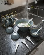 A complete 15-piece ceramic soup set displayed in a kitchen setting, featuring one large scalloped serving bowl, six small individual soup bowls, a golden metal storage stand, and seven white serving spoons. The set is available in six colors: black, dark gray-blue, navy blue, emerald green, beige/nude, and light mint green, all highlighted with an elegant gold rim.