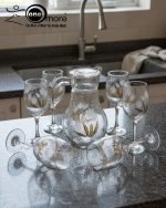 7-piece glass water set, including a clear glass jug/pitcher with white lid and six stemmed wine glasses, all featuring an elegant gold floral design, shown serving beverages in a home setting.