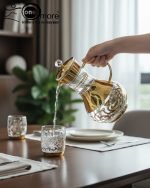 Elegant 7-piece glass beverage set with gold accents including a decanter and six tumblers, shown on a table setting and being used to pour a golden drink.