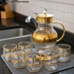 Elegant 7-piece glass beverage set with gold accents including a decanter and six tumblers, shown on a table setting and being used to pour a golden drink.
