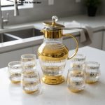 Elegant 7-piece glass beverage set with gold accents including a decanter and six tumblers, shown on a table setting and being used to pour a golden drink.
