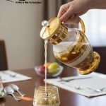 Elegant 7-piece glass beverage set with gold accents including a decanter and six tumblers, shown on a table setting and being used to pour a golden drink.