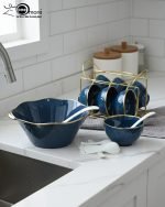 A complete 15-piece ceramic soup set displayed in a kitchen setting, featuring one large scalloped serving bowl, six small individual soup bowls, a golden metal storage stand, and seven white serving spoons. The set is available in six colors: black, dark gray-blue, navy blue, emerald green, beige/nude, and light mint green, all highlighted with an elegant gold rim.