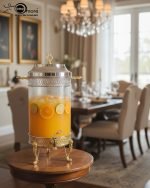 Luxury stainless steel and clear glass beverage dispenser with ornate gold-tone base and handles, available in 7-liter and 10-liter sizes, shown serving infused water at an elegant event.
