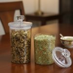 Clear acrylic storage jars with gold leaf pattern, featuring one tall and one short jar, shown filled with colorful Indian spices/mukhwas on a wooden table. Jars have matching clear acrylic lids with round knob handles.
