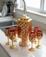 Fancy gold-tone beverage dispenser set with six matching stemmed goblets. The dispenser and glasses have a repeating geometric lattice pattern available in white and gold, blue and gold, or red and gold.