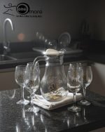 7-piece glass water set, including a clear glass jug/pitcher with white lid and six stemmed wine glasses, all featuring an elegant gold floral design, shown serving beverages in a home setting.