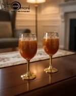 Fancy gold-tone beverage dispenser set with six matching stemmed goblets. The dispenser and glasses have a clear body overlaid with a delicate, scrolling gold floral pattern, and the goblets have solid gold bases.