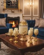 Fancy gold-tone beverage dispenser set with six matching stemmed goblets. The dispenser and glasses have a clear body overlaid with a delicate, scrolling gold floral pattern, and the goblets have solid gold bases.