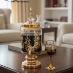 Fancy gold-tone beverage dispenser set with six matching stemmed goblets. The dispenser and glasses have a clear body overlaid with a delicate, scrolling gold floral pattern, and the goblets have solid gold bases.