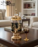Fancy gold-tone beverage dispenser set with six matching stemmed goblets. The dispenser and glasses have a clear body overlaid with a delicate, scrolling gold floral pattern, and the goblets have solid gold bases.