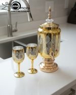 Fancy gold-tone beverage dispenser set with six matching stemmed goblets. The dispenser and glasses have a clear body overlaid with a delicate, scrolling gold floral pattern, and the goblets have solid gold bases.
