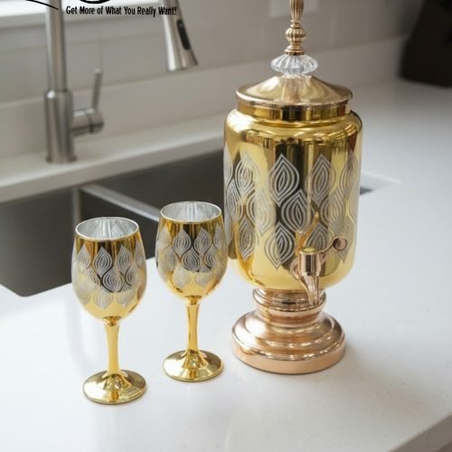 Fancy gold-tone beverage dispenser set with six matching stemmed goblets. The dispenser and glasses have a clear body overlaid with a delicate, scrolling gold floral pattern, and the goblets have solid gold bases.