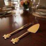 Luxurious golden cake knife and serving set with detailed palm leaf handles resting on a polished dark wood dining table, set for a formal dinner.