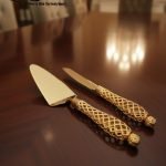 Close-up of a luxurious gold cake knife and matching server set with intricately detailed lattice handles, resting on a polished mahogany table. The scene features cinematic lightening (CL) and a blurred background (BG) of a dining room.