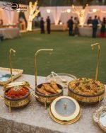 A collection of luxurious gold and deep brown Royal Buffet Server Chafing Dishes, available in 1.5, 2.2, and 3.2-liter sizes. The dishes feature intricate lattice detailing, clear-top lids, and elegant gold stands with hooks for convenient lid placement. The images show the servers beautifully displayed on formal dining and outdoor buffet tables, holding hot dishes like Biryani and curry, emphasizing their elegant design and food-warming capability for weddings and upscale events.
