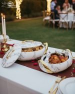 A set of three stacked white marble-patterned insulated casserole dishes (hot pots) with striking gold-tone handles and lids, displayed on a formal dining table.