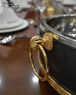 Set of three black insulated hot pot casseroles with gold trim and loop handles, sitting on a wooden dining table with a formal place setting.
