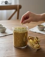 A set of three ribbed acrylic storage jars in different sizes, some with clear lids and some with shiny gold lids, filled with various nuts and snacks, displayed on a wooden table.