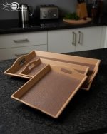 Set of three nesting fiber serving trays with non-slip textured surfaces and cut-out handles, displayed on a wooden dining table. The available colors are black, dark brown, grey, off-white, and rust-brown.