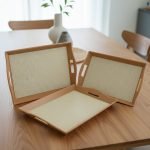 Set of three nesting fiber serving trays with non-slip textured surfaces and cut-out handles, displayed on a wooden dining table. The available colors are black, dark brown, grey, off-white, and rust-brown.