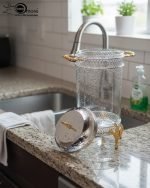 Luxury stainless steel and clear glass beverage dispenser with ornate gold-tone base and handles, available in 7-liter and 10-liter sizes, shown serving infused water at an elegant event.