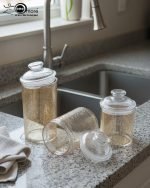 A set of three ribbed acrylic storage jars in different sizes, some with clear lids and some with shiny gold lids, filled with various nuts and snacks, displayed on a wooden table.