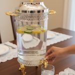 Luxury stainless steel and clear glass beverage dispenser with ornate gold-tone base and handles, available in 7-liter and 10-liter sizes, shown serving infused water at an elegant event.